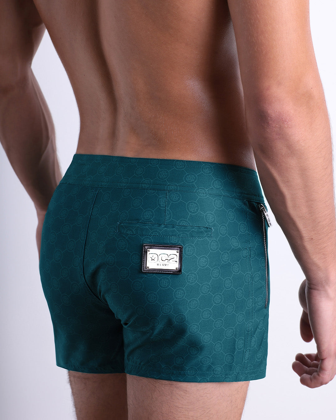 Back view of a male model wearing men’s Summer MONO TEAL Beach Shorts in a teal color with a lighter blue monogram logo, complete with a back pocket, designed by DC2. made by DC2 a capsule brand by BANG! Clothes in Miami.