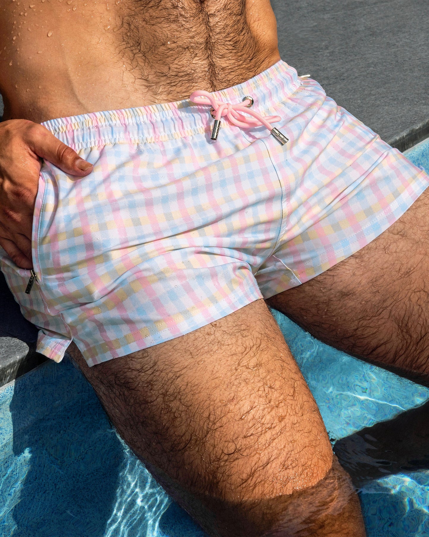 Close-up detail of the Ocean Drive Check Poolside Shorts featuring a pastel plaid print in soft pink, lavender, cream, and sky blue tones beside a luxury pool setting.