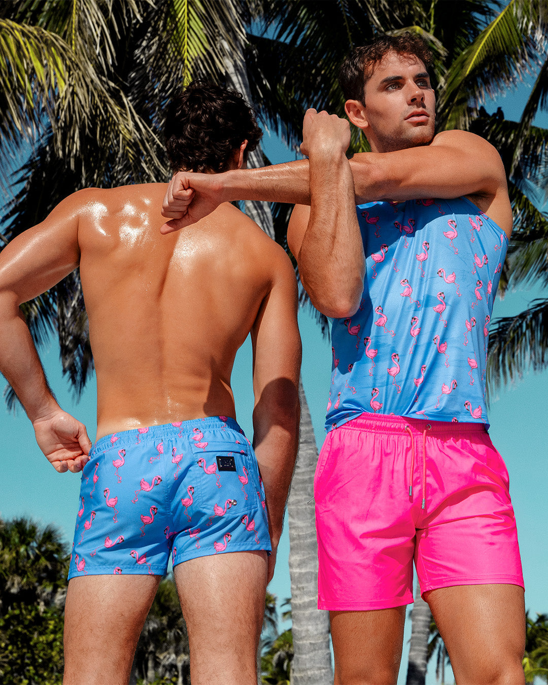 Two male models in tropical setting wearing DC2 Miami summer outfits. Left model wears PINK FLAMINGO Poolside Shorts in blue with pink flamingo print. Right model wears PINK BOMB Resort Shorts in hot pink and matching flamingo print tank top.