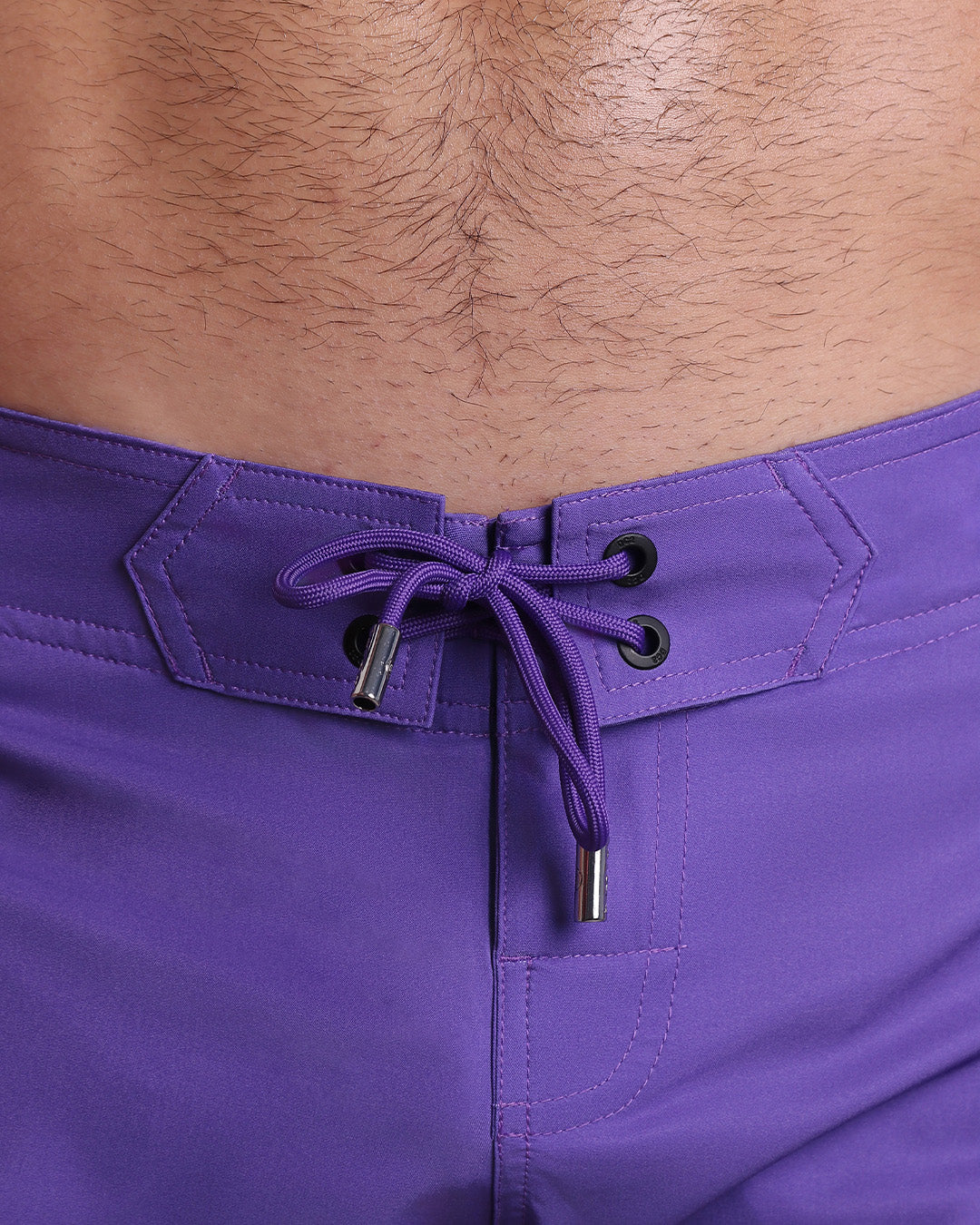 Close-up view of inseam and details of PURPLE MACHINE beach shorts for men, with a purple color cord and custom branded silver cord-ends, and matching custom eyelet trims in black.