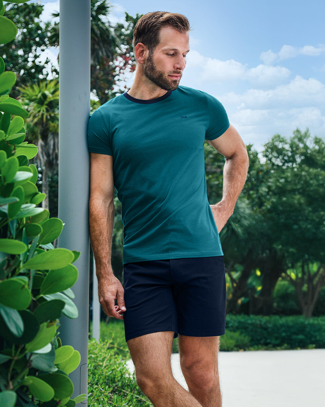 Model wearing DC2 Ring On It Teal Pima Cotton T-Shirt and Universal Blue Street Shorts, leaning casually outdoors, showcasing a sleek and modern resort-inspired look.