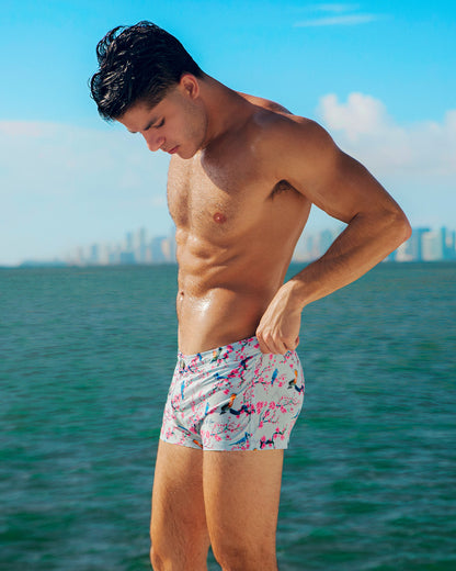 This image features a model wearing CLOSE TO YOU Mini Shorts, with the Miami skyline in the backdrop. The beach shorts have an exotic bird design in light blue color with pink flowers, designed for men. These premium quality swimwear bottoms are by DC2, a popular men’s beachwear brand based in Miami.