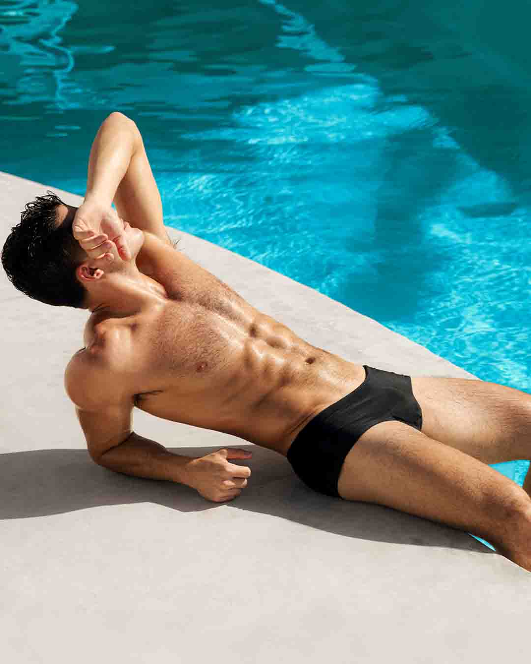 Sexy model under the sun in swimming pool wearing the Swim Brief silhouette of the new JET BLACK dark color men's swimsuit by DC2 Miami for BANG menswear Miami.