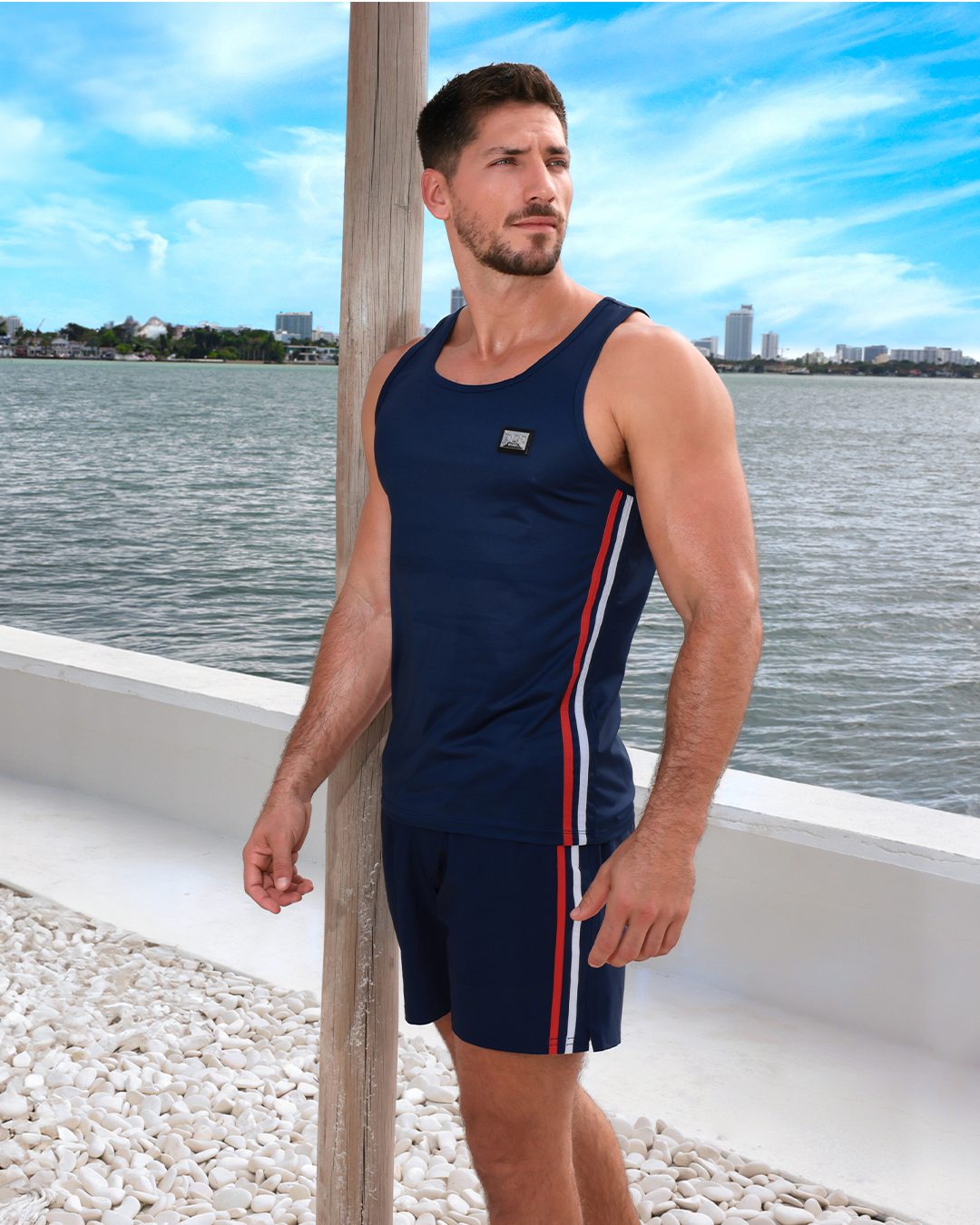 Fit male model wearing IMPERIAL BLUE Resort Shorts and navy blue tank top, leaning on a wooden post by the water with a clear blue sky overhead