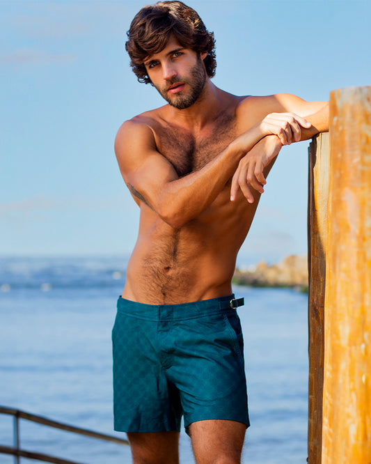 Male model leaning against a wooden post wearing MONO TEAL Tailored Shorts by DC2 Miami. These stylish men's shorts feature a subtle geometric pattern and a tailored fit, perfect for beachwear and resort fashion.