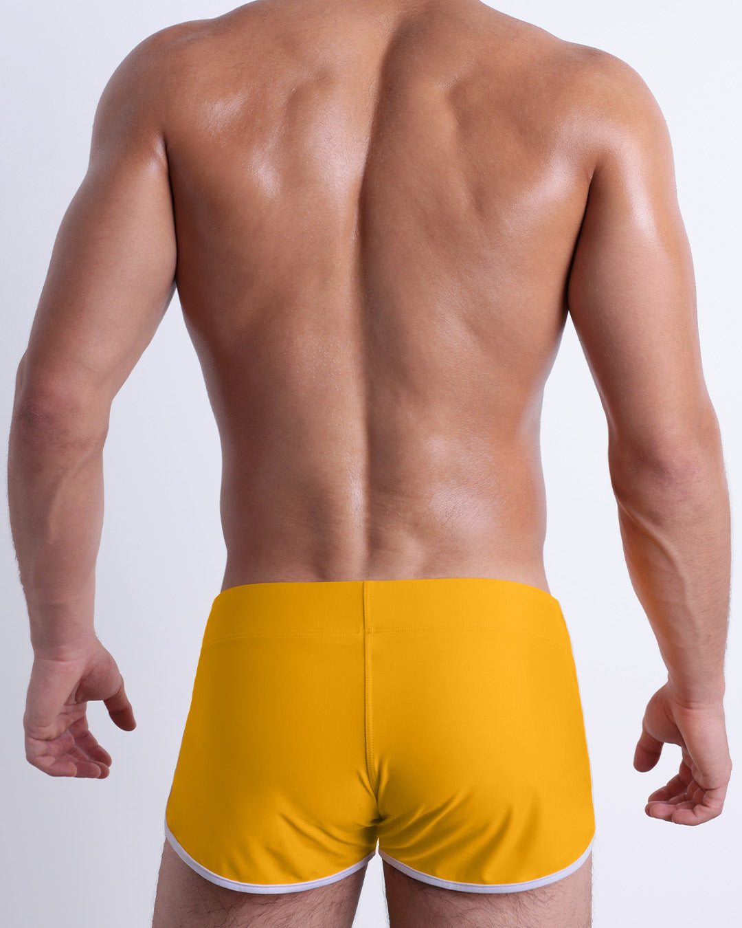 Back view of a male model wearing the AMBER SAND beach Swim Shorts for men by BANG! Miami in a solid dark yellow color.