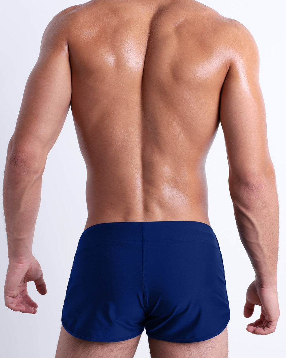 Back view of male model wearing the BLUE BY THE OCEAN beach Swim Shorts for men by BANG! Miami in a solid dark blue color.