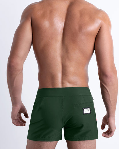 Back view of a male model wearing men’s Summer BRAVE GREEN Beach Shorts in a solid dark green color with orange and white side stripes, complete the back pockets, made by DC2 a capsule brand by BANG! Clothes in Miami.