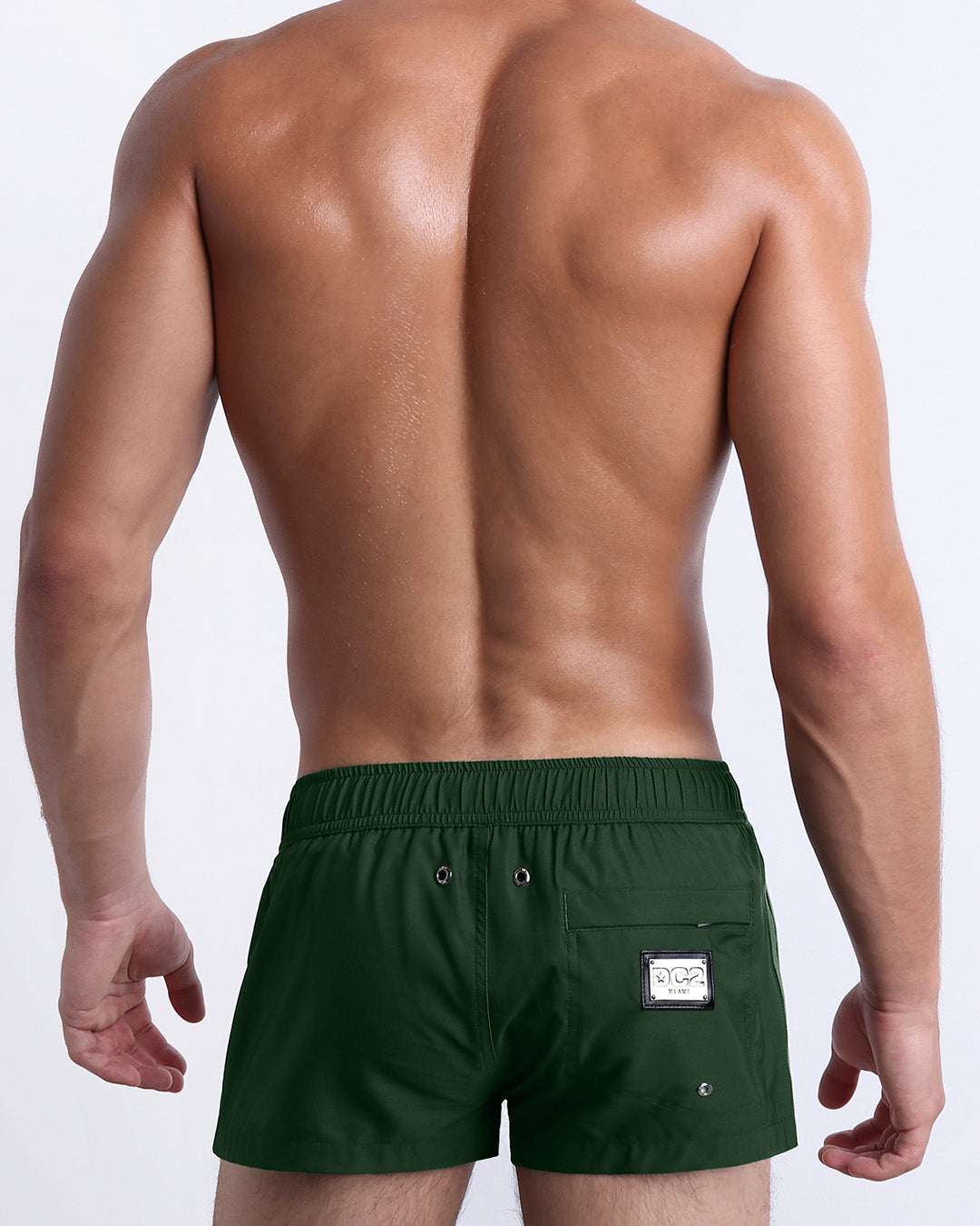 Back view of male model wearing men’s BRAVE GREEN beach Mini Shorts swimsuits in a solid green color with side stripes in orange and white colors, complete with a back pocket, designed by DC2 a capsule brand by BANG! Clothes based in Miami.