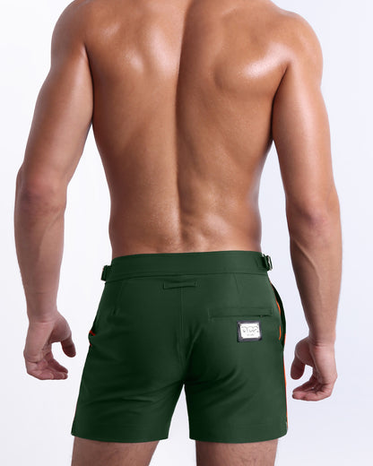 Male model wearing men’s BRAVE GREEN beach Tailored Shorts swimsuit in a solid green color with side stripes in orange and white colors, complete with a back pocket, designed by DC2 a capsule brand by BANG! Clothes based in Miami.