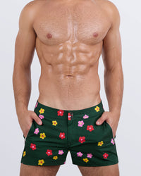 Frontal view of a masculine model wearing the BUY MYSELF FLOWERS beach shorts for men by DC2 Miami, a premium swimwear brand. These modern-fit swimming shorts feature a deep green base with vibrant red, pink, and yellow floral prints, offering a bold, stylish look perfect for beach days, poolside lounging, and summer adventures.