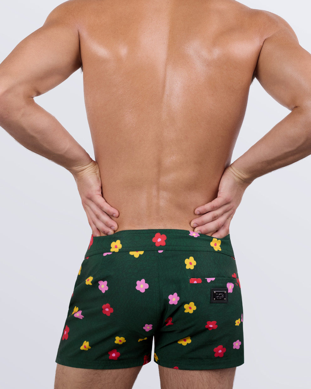 Back view of a male model wearing the men's BUY MYSELF FLOWERS beach shorts by DC2 Miami. Featuring a playful floral pattern on a rich green backdrop, a sleek fit, and a functional back pocket with a DC2 signature metal logo plaque, these premium swim shorts combine eye-catching design with ultimate comfort.