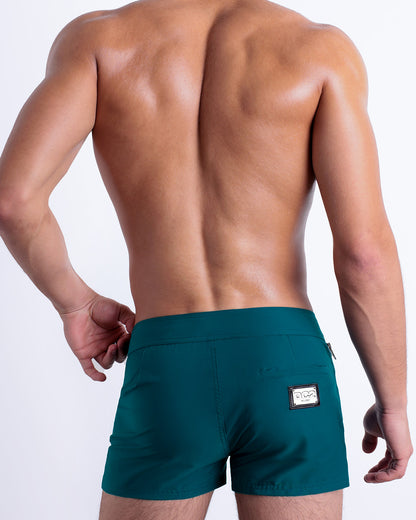 Male model wearing men’s CHIC TEAL Beach Shorts swimsuit in a dark aqua green blue color, complete the back zippered pocket, made by DC2 a capsule brand by BANG! Clothes in Miami.