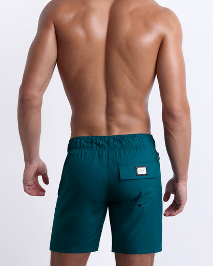 Back view of male model wearing men’s CHIC TEAL beach Flex Boardshorts swimming shorts. In a solid dark teal color, complete with a back pocket, designed by DC2 a capsule brand by BANG! Clothes based in Miami.