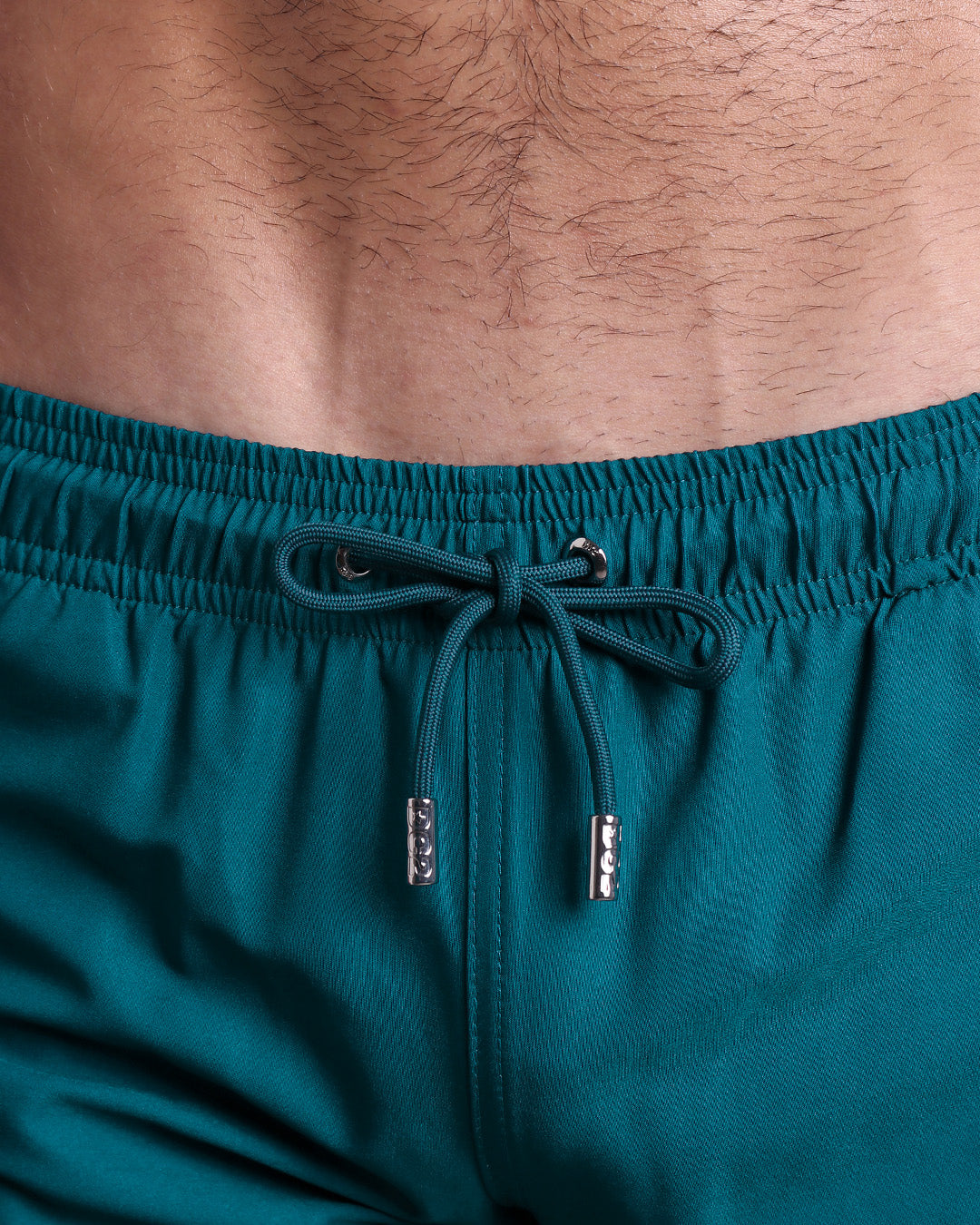 Close-up view of the CHIC TEAL men’s summer shorts, showing blue green cord with custom branded silver cord ends, and matching custom eyelet trims in silver.