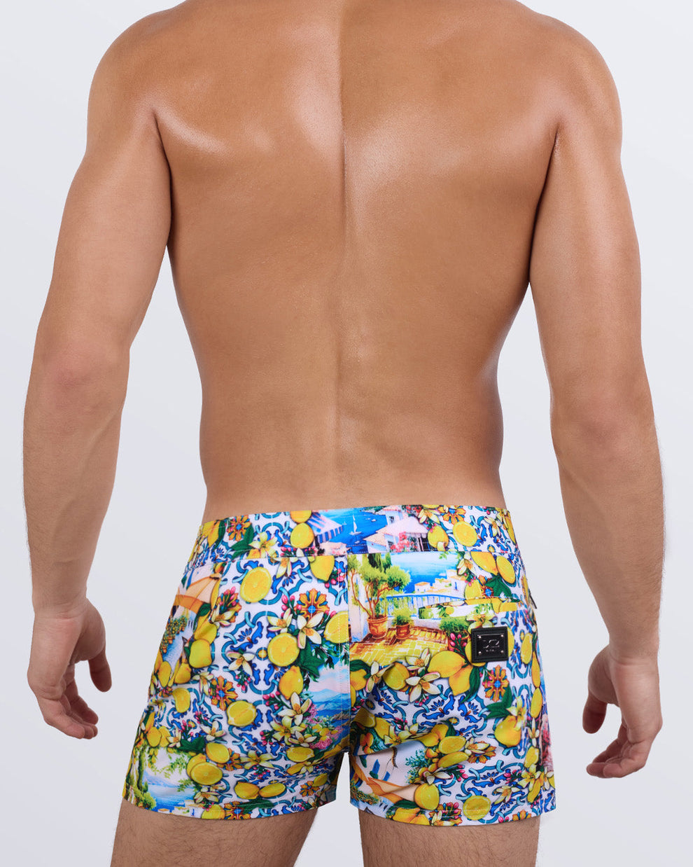 Back view of a male model wearing the men's CITRICS CHOICE beach shorts by DC2 Miami. Featuring a stunning lemon and floral print reminiscent of a Mediterranean summer, a sleek fit, and a functional back pocket with a DC2 signature metal logo plaque, these premium swim shorts combine eye-catching design with ultimate comfort.