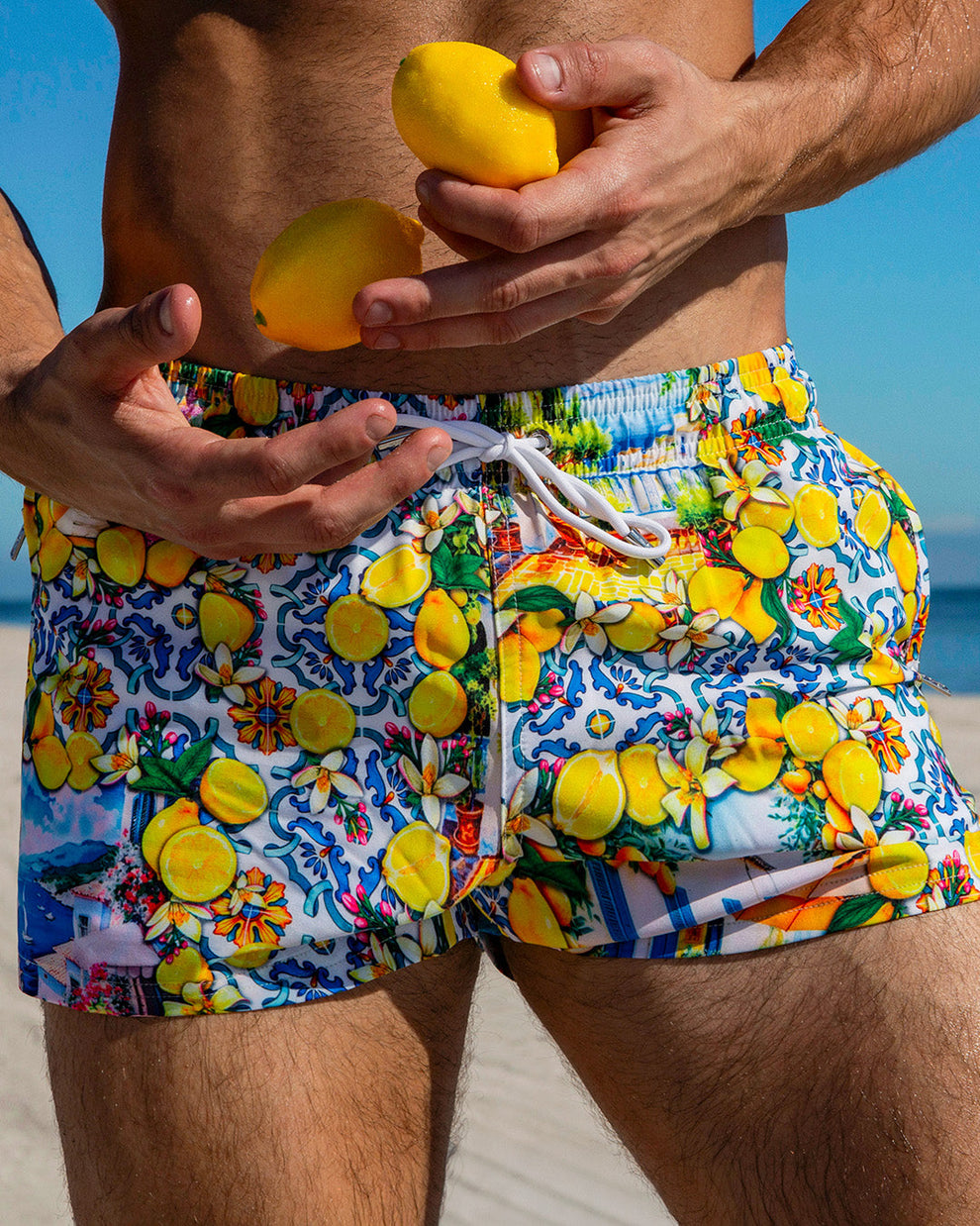 Close-up of male model wearing DC2 Miami Citrics Choice men's poolside swim shorts with vibrant Amalfi Coast-inspired lemon and tile print, holding two lemons in hand at the beach.