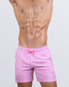 Frontal view of a male model wearing the FAST LINES (CHILL PINK) Resort Shorts for men by DC2 Miami. These above-the-knee swim shorts feature a stylish pink and white striped pattern, an elastic waistband with an adjustable drawstring, and a relaxed fit, perfect for poolside lounging, tropical vacations, and summer getaways.