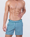 Model wearing FAST LINES (PACIFIC GREEN) tailored swim shorts for men with bold vertical stripes in green and white. High-end men’s swim trunks with a slim fit, adjustable side buckles, and quick-dry fabric. Perfect for beach days and resort fashion.