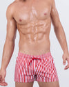 Model wearing FAST LINES (RAPID RED) poolside shorts in bold red and white vertical stripes. Athletic fit swimwear with adjustable drawstring waistband, made for standout beach looks.
