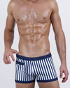Model wearing FAST LINES (NAVY BLUE) men's swim trunks with vertical navy and white stripes. Features a square-cut silhouette, skin-hugging fit, and front mini zipper pocket for functional summer style.