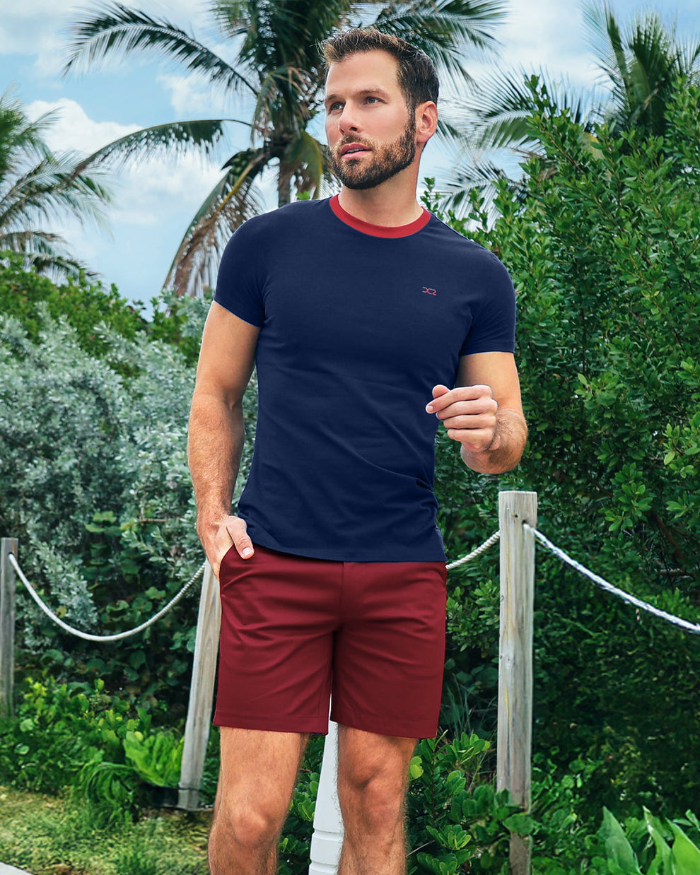 Model wearing DC2 Fontaine Blue Pima Cotton T-Shirt and Under My Umbrella Street Shorts, walking along a tropical path, showcasing a bold navy and red resort look.