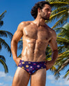 DC2 Flower Powers Blue Swim Brief — navy swim brief featuring colorful scattered floral prints, styled under tropical palm trees.