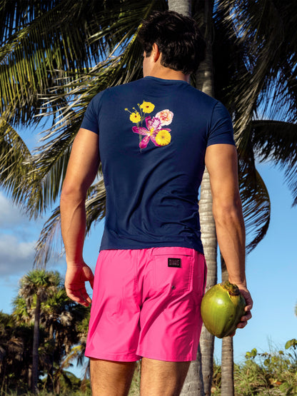 Model wearing Pink Bomb Resort Shorts with the Flower Powers Blue Graphic Cotton T-Shirt, shown from the back on a tropical beach, highlighting the floral back graphic, bright pink swim shorts and relaxed resort style.