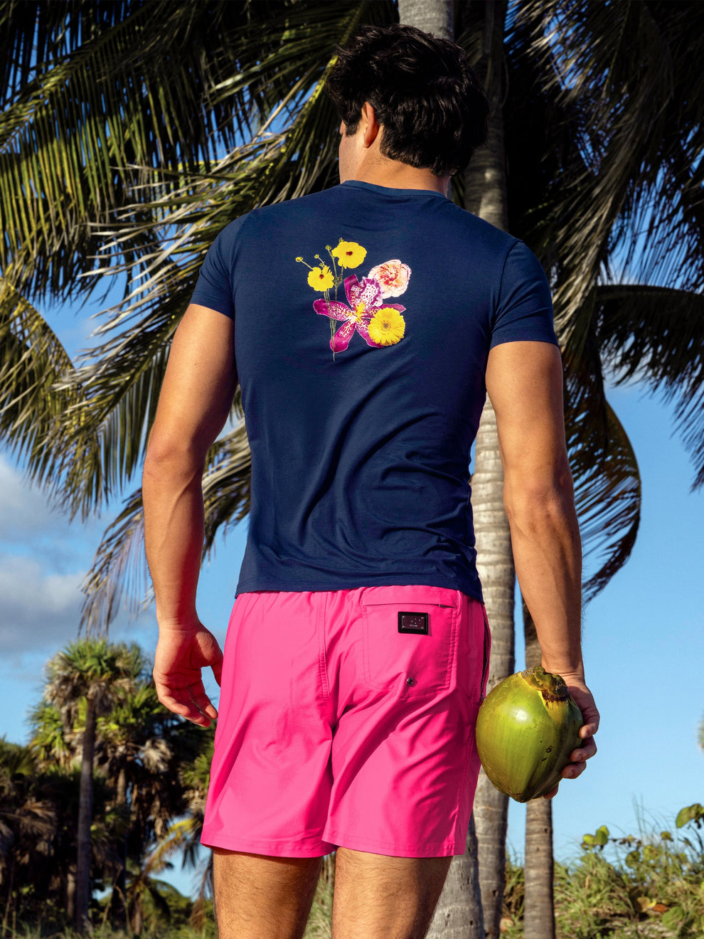 Model wearing Pink Bomb Resort Shorts with the Flower Powers Blue Graphic Cotton T-Shirt, shown from the back on a tropical beach, highlighting the floral back graphic, bright pink swim shorts and relaxed resort style.