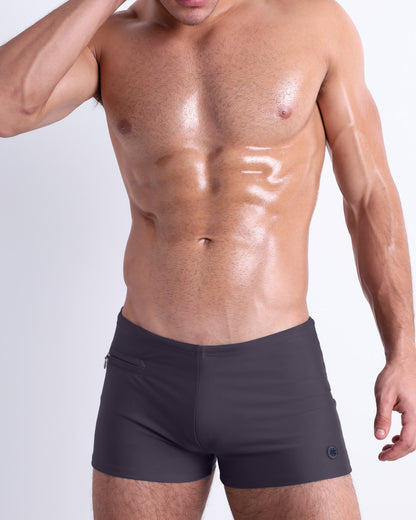 Frontal view of a sexy male model wearing men’s swimsuit with mini pockets in a solid dark grey color by DC2, a men's beachwear brand from Miami.