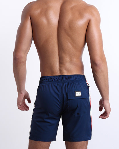 Back view of a male model wearing men’s IMPERIAL BLUE Flex Boardshorts swimsuits in a solid navy blue color with white and red side stripes, complete the back pockets, made by DC2 a capsule brand by BANG! Clothes in Miami.