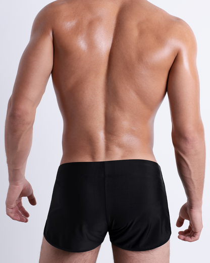 Back view of a male model wearing the JET BLACK beach Swim Shorts for men by BANG! Miami in a solid black color.