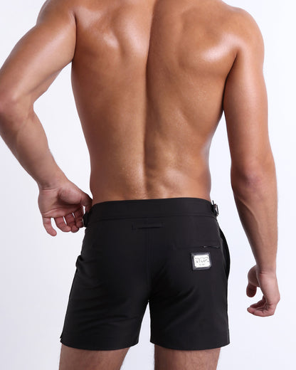 Male model wearing men’s JET BLACK Summer Tailored Shorts swimsuit in a solid black color, complete with a back zippered pocket, designed by BANG! Clothes in Miami.