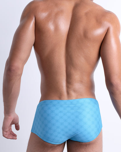 Back view of male model wearing the MONO BLUE beach sexy Brazilian Sunga for men, with a stylish DC2 logo monogram motif in a blue print, designed by DC2.