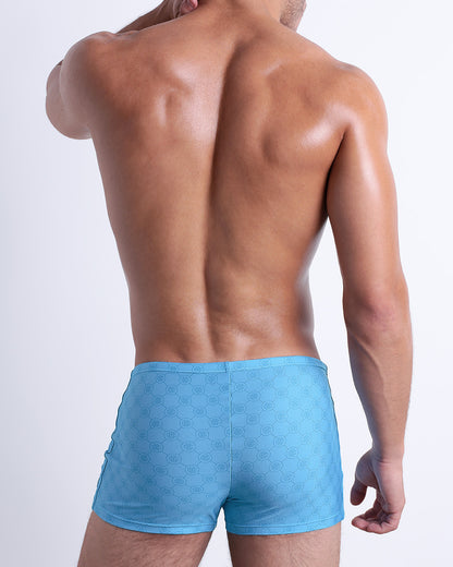 Back view of male model wearing the MONO BLUE beach sexy swimming bottoms for men in a blue color with a blue monogram logo, designed by DC2.