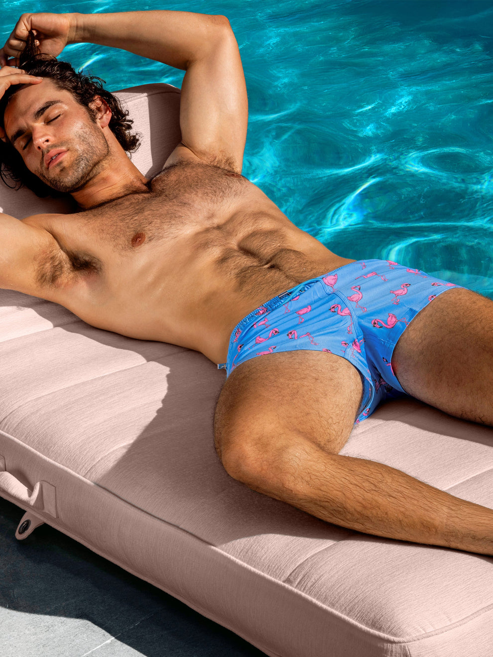 Model lounging poolside wearing the Flamingo Blue Poolside Shorts, featuring a playful pink flamingo print on a bright blue background.