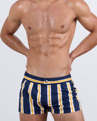 Frontal view of a masculine model wearing the SUN CHASING STRIPES Mini Shorts for men by DC2 Miami, a premium swimwear brand. These ultra-short beach shorts feature a bold navy blue base with striking yellow and white stripes, an elastic waistband with an adjustable internal drawstring, and a sleek fitted cut, making them perfect for stylish summer looks, beach days, and poolside lounging.