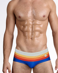 DC2 Sunset Stripes — model wearing Sunset Stripes Swim Sunga featuring a bold royal blue base with layered horizontal stripes in yellow, orange, and red across a soft sand-toned waistband.