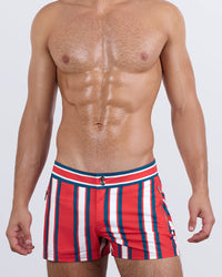 Model wearing SWIFT STRIPES men's beach shorts in a bold red base with teal and white vertical stripes. Features include a flat waistband, dual button closure, and zippered side pocket for secure storage.