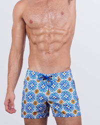 A male model wearing THE TILE IS HIGH - Flex Shorts by DC2 Miami. These premium swim shorts feature a striking blue and yellow tile-inspired pattern, a lace-up front, and water-resistant metallic zippers.