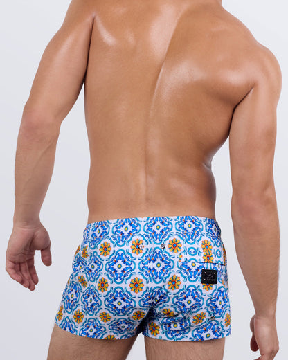 Back view of a male model wearing the men's THE TILE IS HIGH Mini Shorts by DC2 Miami. Featuring a striking Mediterranean tile pattern, a tailored ultra-short fit, and a functional back pocket with a DC2 signature metal logo plaque, these stylish swim shorts offer both comfort and elegance for beach lovers.