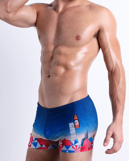 Side view of a muscular male model wearing UNDER MY UMBRELLA Summer Swimming Trunks. Featuring a photorealistic beach scene print designed by DC2 a brand based in Miami.