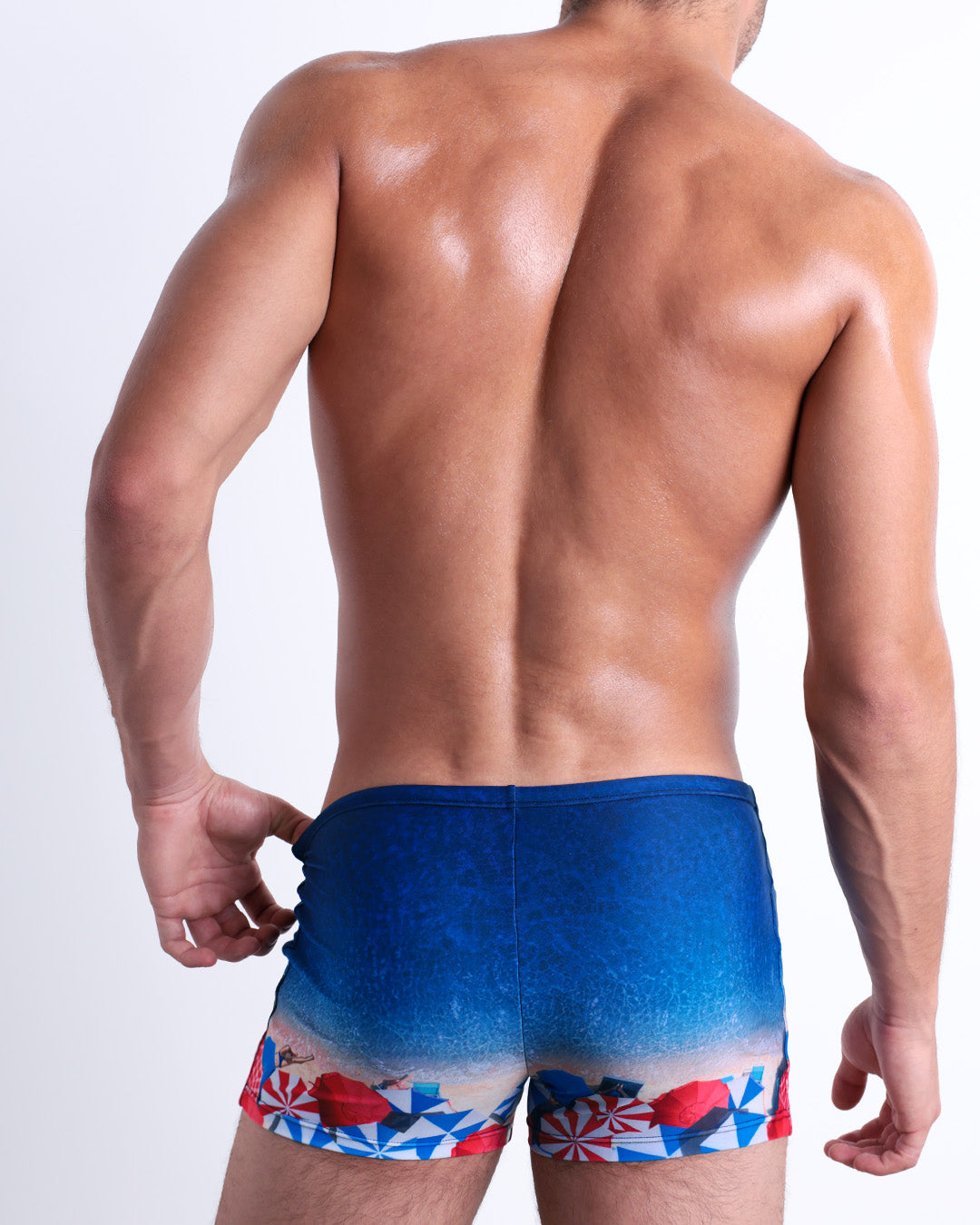 Back view of male model wearing the UNDER MY UMBRELLA beach sexy swimming bottoms. Featuring a photorealistic beach scene print designed by DC2 a brand based in Miami.